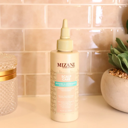 Une bouteille de spray Mizani Scalp Care Reviving & Refreshing est posée sur le comptoir d'une salle de bain. L'arrière-plan présente un mur carrelé beige et une petite plante dans un pot blanc. La bouteille est dotée d'un bouchon à bec pointu. Dakar